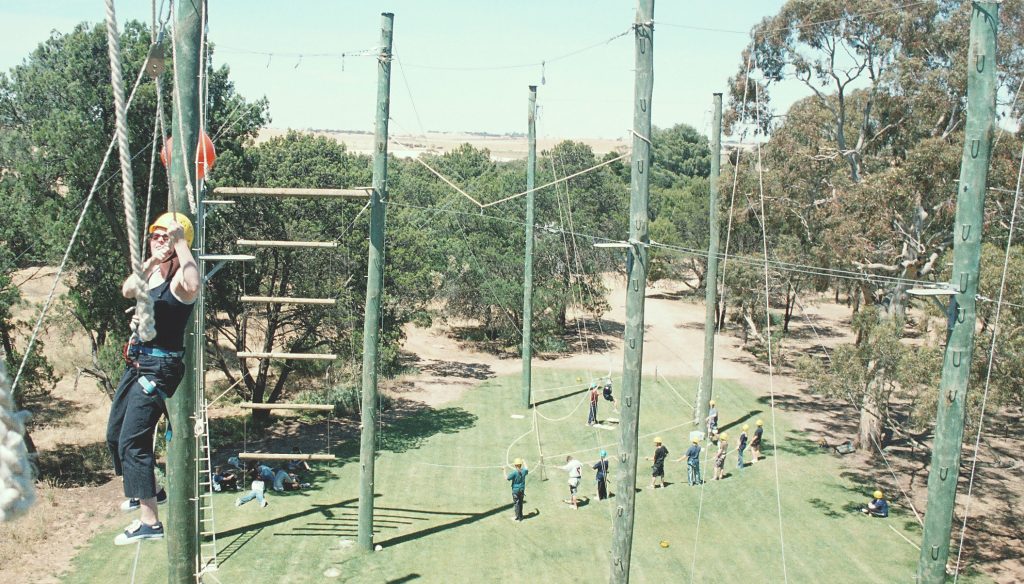 Glenhaven Park Camps A person wearing a helmet and harness climbs a high ropes course while a group of people waits below, creating an adventurous scene perfect for your outdoor activity company's home landing page.