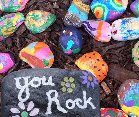 Glenhaven Park Camps A collection of colorful, painted rocks on the ground. The largest rock in the foreground has the words "You Rock" painted on it along with flower designs, showcasing creativity from recent camp craft activities during a memorable campout.