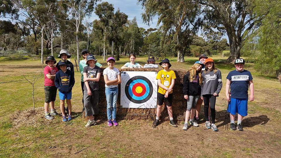 Glenhaven Park Camps A group of children stand outdoors in front of an archery target, some wearing baseball caps and hats. Trees and greenery can be seen in the background. Such school camps foster not only fun but also contribute to teachers' growth and the wellbeing of students.