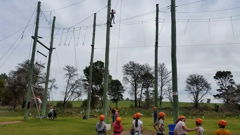 Glenhaven Park Camps People wearing helmets participate in a high ropes course outdoors on a grassy field, with some climbing and others watching from below. This activity, often featured in school camps, fosters not only adventure but also supports teachers' growth and wellbeing.